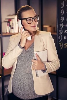 Portrait of a young beautiful pregnant female in the office, expectant business woman talking on the phone with a business partner, active pregnancy concept