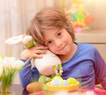 Portrait of cute little boy with Easter bunny toy sitting on the kitchen near colorful eggs, traditional festive food, preparation for great religious holiday