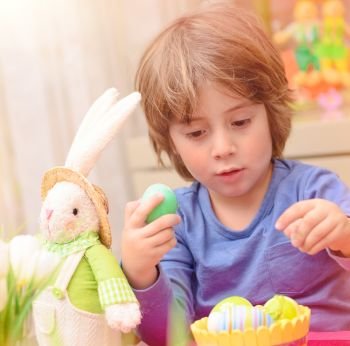 Portrait of cute little boy with Easter bunny toy sitting on the kitchen and coloring eggs, traditional festive food, preparation for great religious holiday