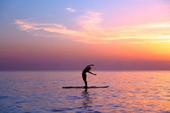 Silhouette of a yoga trainer over sunset background doing asanas, balancing on the sup board, enjoying healthy lifestyle, summer vacation on the beach
