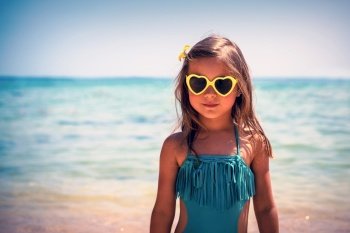 Portrait of a cute little girl on the beach, charming child wearing stylish swimsuit and heart shaped sunglasses, baby fashion, happy summer holidays
