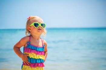 Cute little blond girl on the beach wearing sunglasses and stylish colorful swimsuit, child’s fashion, summer vacation near the sea