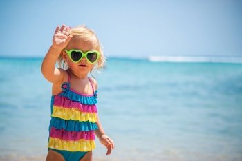 Cute little blond girl on the beach wearing sunglasses and stylish colorful swimsuit, child’s fashion, smalll model posing near seashore
