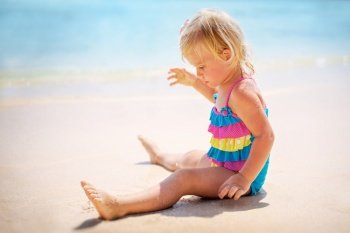 Cute little baby girl on the beach, with pleasure sitting on the seashore and playing in sand, happy summer holidays near the sea
