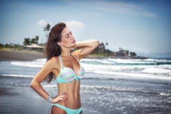 Hot woman standing on a beach and with closed eyes enjoying nice sea breeze, girl model relaxing on a tropical spa resort, peaceful summer vacation