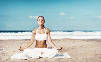 Pretty woman with pleasure meditating outdoors, sitting on the beach in lotus pose with closed eyes and doing yoga asana, peace and zen balance concept