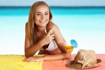 Portrait of a pretty smiling female with pleasure spending time on the beach,  uses a sunscreen spray for a safe tan, applying anti-aging moisturizing cream for staying under the sun, healthy tan