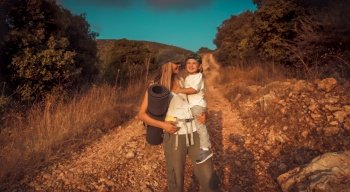 Happy traveler family, active young mother with cute little son on hands walking along mountainous road, enjoying summer camping in the mountains