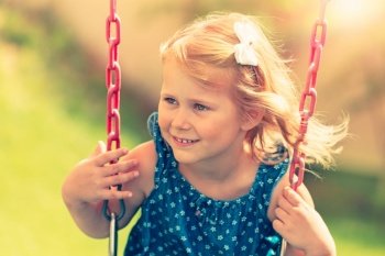 Portrait of a cute little blond girl on a swing on playground outdoors, sweet child with pleasure spending summer weekend in the park