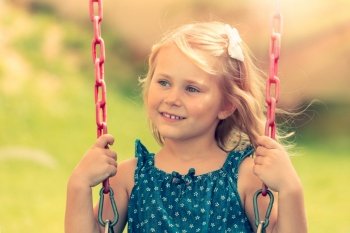 Portrait of a cute little blond girl riding on a swing on a playground outdoors, sweet child with pleasure spending summer weekend in the park