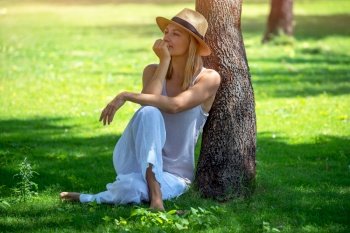 Pretty Woman in the Park. Nice Calm Female Sitting on the Ground near Tree Trunk on Sunny Green Grass Glade. Enjoying Mild Spring Summer Weather.. Pretty Woman in the Park