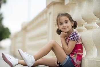 Adorable little girl combed with pigtails outdoors sitting on urban floor.