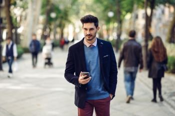 Attractive man in the street wearing british elegant suit with smart phone in his hand. Young bearded businessman with modern hairstyle in urban background.