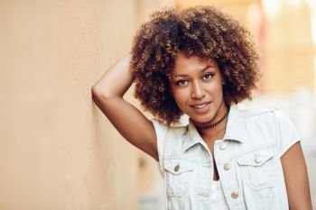 Young black woman, afro hairstyle, smiling near a wall in the street. Girl wearing casual clothes in urban background.