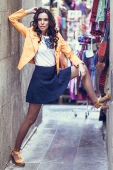 Young brunette woman, model of fashion, wearing orange modern jacket and blue skirt,  in urban background.