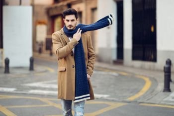 Young man wearing winter clothes in the street. Young bearded guy with modern hairstyle with coat, scarf, blue jeans and t-shirt.