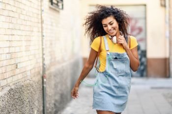 Young African woman with headphones and black curly hairstyle walking outdoors. Happy girl wearing yellow t-shirt and denim dress in urban background.