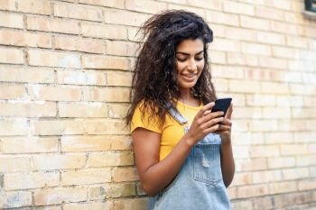 Happy Arab girl using smart phone on brick wall. Smiling woman with curly hairstyle in casual clothes in urban background.