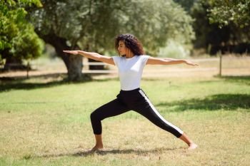 Young Arab woman doing yoga in nature. North African female wearing sport clothes doing Warrior II figure in urban park.