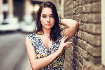 Girl with blue eyes standing next to brick wall outdoors. Young woman in her twenties wearing flower dress in urban background. Beauty and fashion concept.