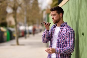 Young man recording voice note in his smart phone in urban background. Guy wearing casual clothes. Lifestyle concept.