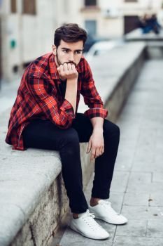 Young bearded man, model of fashion, sitting in urban background wearing casual clothes. Guy with beard and modern hairstyle in the street with plaid shirt.
