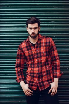 Young bearded man, model of fashion, wearing a plaid shirt with a green blind behind him. Guy with beard and modern hairstyle in urban background.