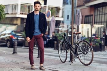 Attractive man in the street wearing british elegant suit near a vintage bicycle. Young bearded businessman with modern hairstyle in urban background.