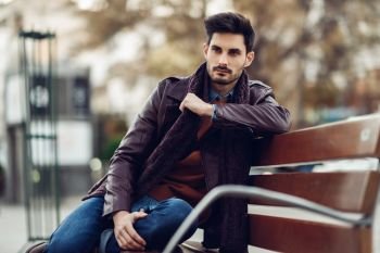 Thoughtful young man sitting on an urban bench. Attractive guy with modern hairstyle with lost look in the street.