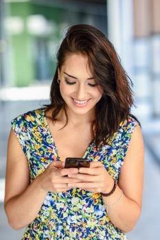 Smiling young woman typing in smart phone outdoors. Girl wearing flower dress in urban background. Technology concept.