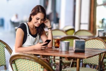 Girl with blue eyes sitting on urban cafe using smart phone smiling. Happy woman with brown wavy hairstyle wearing white denim dress. Lifestyle concept.