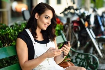 Smiling young woman looking at smart phone outdoors. Girl wearing white denim dress sitting in urban bench. Technology concept.