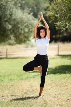 Young Arab woman doing yoga in nature. North African female wearing sport clothes doing tree figure in urban park.