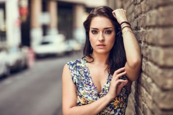 Beautiful young woman with blue eyes standing next to brick wall outdoors. Girl wearing flower dress in urban background. Beauty and fashion concept.