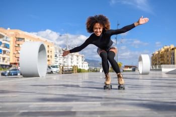 Young fit black woman on roller skates riding outdoors on urban street with open arms. Smiling girl with afro hairstyle rollerblading on sunny day