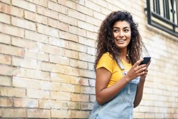 Happy Arab girl using smart phone on brick wall. Smiling woman with curly hairstyle in casual clothes in urban background.