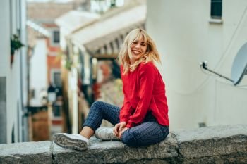Happy young blond woman sitting on urban background moving her hair. Smiling blonde girl with red shirt enjoying life outdoors.