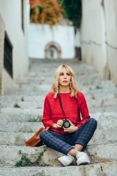 Attractive young blond woman taking photographs with an old slr camera in a beautiful city. Blonde happy woman sitting on urban steps.