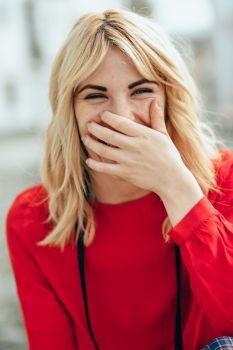 Happy young blond woman sitting on urban background laughing. Smiling blonde girl with red shirt enjoying life outdoors.