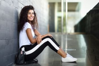 Happy African girl with black curly hairstyle sitting on urban floor. Smiling Arab woman in sport clothes in the street.
