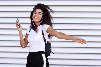 Funny African woman listening to music and dancing with earphones and smartphone outdoors. Arab girl in sport clothes with curly hairstyle in urban background.