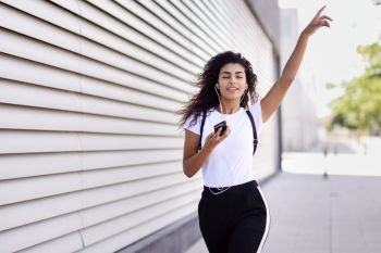 Funny African woman listening to music and dancing with earphones and smartphone outdoors. Arab girl in sport clothes with curly hairstyle in urban background.. African woman listening to music with earphones and smartphone