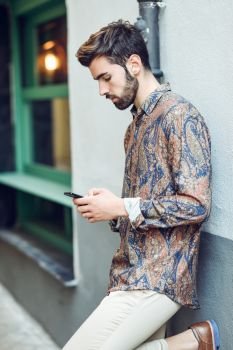 Young man wearing casual clothes looking at his smartphone in the street. Guy with beard and modern hairstyle in urban background. Young man wearing casual clothes looking at his smartphone in the street.
