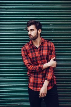 Young bearded man, model of fashion, wearing a plaid shirt with a green blind behind him. Guy with beard and modern hairstyle in urban background.. Young bearded man, model of fashion, wearing a plaid shirt with a green blind behind him. 
