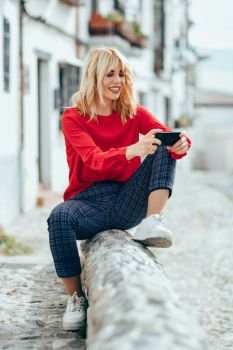 Happy young blond woman sitting on urban background. using smart phone. Smiling blonde girl with red shirt.