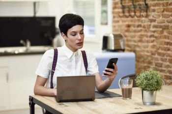 Young woman with very short haircut looking at her smart phone. Businesswoman working at home concept.. Young businesswoman with very short haircut looking at her smartphone at home.