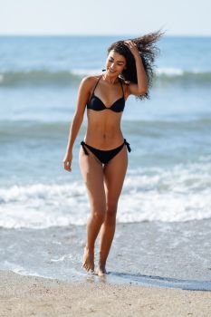 Young arabic woman with beautiful body in swimwear on a tropical beach with moving hair by the wind. Brunette female with curly long hairstyle wearing black bikini.. Young arabic woman with beautiful body in swimwear on a tropical beach.