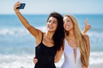 Two young women taking selfie photograph with smart phone in swimsuits on a tropical beach.. Two women taking selfie photograph with smartphone in the beach