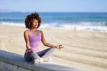 Black woman, afro hairstyle, doing yoga asana in the beach with eyes closed. Young Female wearing sport clothes in lotus pose with defocused background.. Black woman, afro hairstyle, in lotus pose with eyes closed in the beach