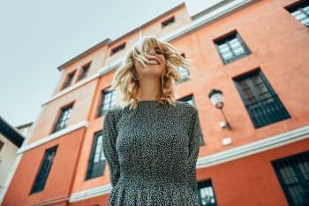 Happy young woman with moving hair in urban background. Blond girl, wavy hairstyle, wearing casual clothes in the street.. Happy young woman with moving hair in urban background.